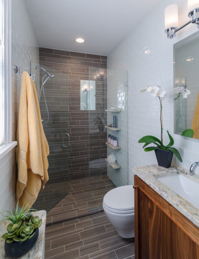 Full-length view of a small bathroom remodel displaying continuous textured stone floor tile running straight into a walk-in glass shower enclosure.