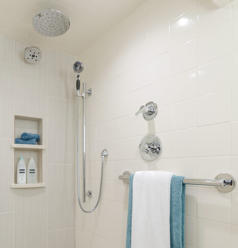 A wide-angle view of a fully remodeled, bright basement wet room bathroom featuring a biscuit-colored tile palette, a classic pedestal sink, a toilet with a floating teak cabinet above, and a barrier-free curbless shower entry enclosed by a seamless glass panel.