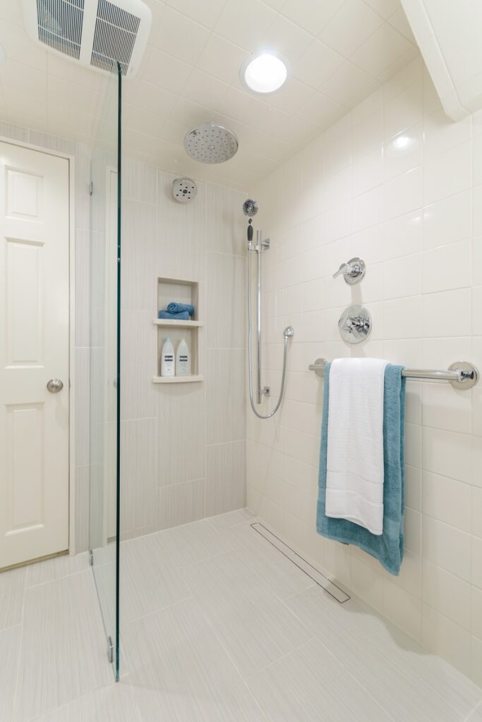 Close-up view of a luxury multi-head shower system in a remodeled wet room, showing stacked chrome plumbing valves, a heavy-duty safety grab bar with a white towel, and a vertical hand-held sprayer against warm biscuit wall tile.