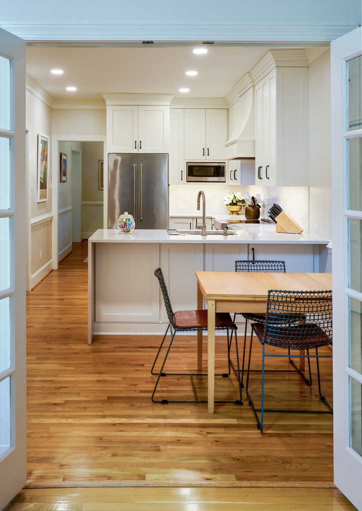 View looking through open French doors into a bright remodeled kitchen featuring white shaker cabinetry, hardwood floors, a quartz peninsula, and a dining table set in the foreground.