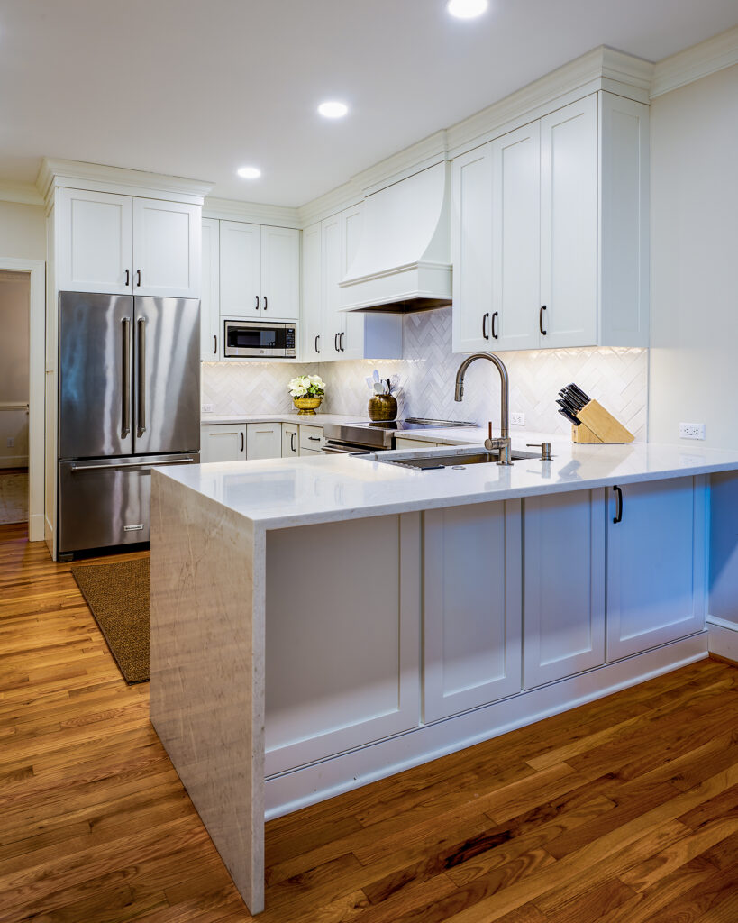 Angle view of a white quartz kitchen peninsula with a waterfall edge, undermount stainless steel sink, Brizo gooseneck faucet, and white shaker upper cabinets