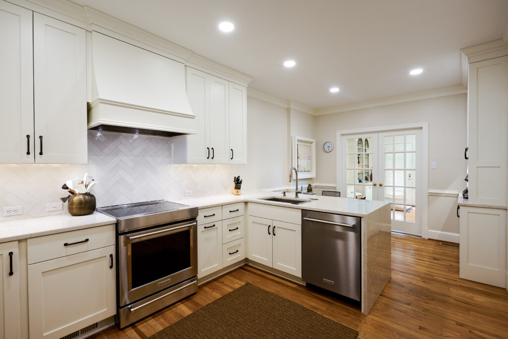 Wide view of the open-concept kitchen highlighting the herringbone ceramic backsplash, custom range hood enclosure, stainless steel dishwasher, and quartz waterfall peninsula.
