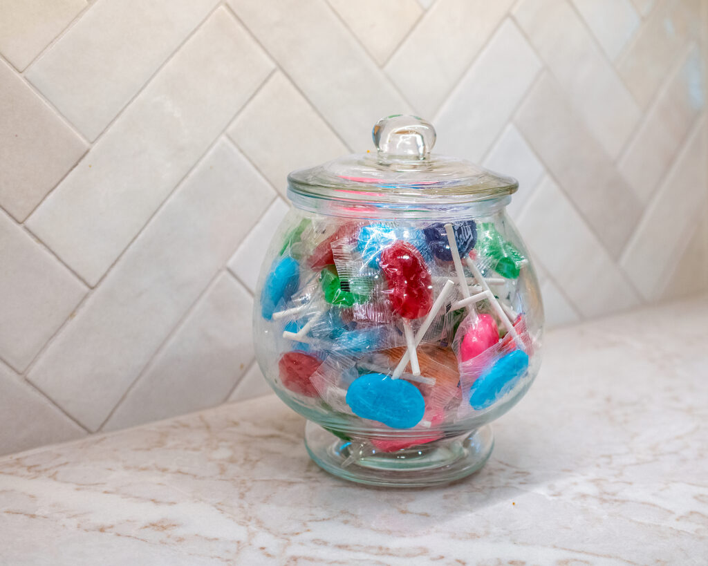 Close-up of a clear glass candy jar filled with colorful lollipops sitting on a Venus quartz countertop against an off-white herringbone tile backsplash.